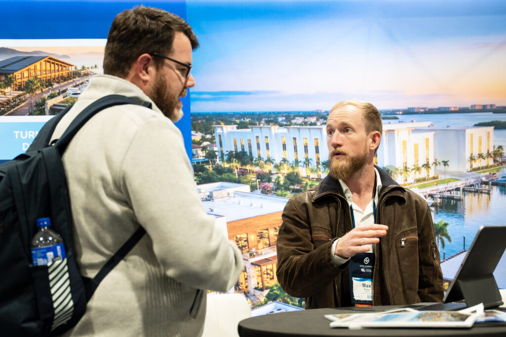 Two men discussing business at a trade show, with various displays and visitors in the background.