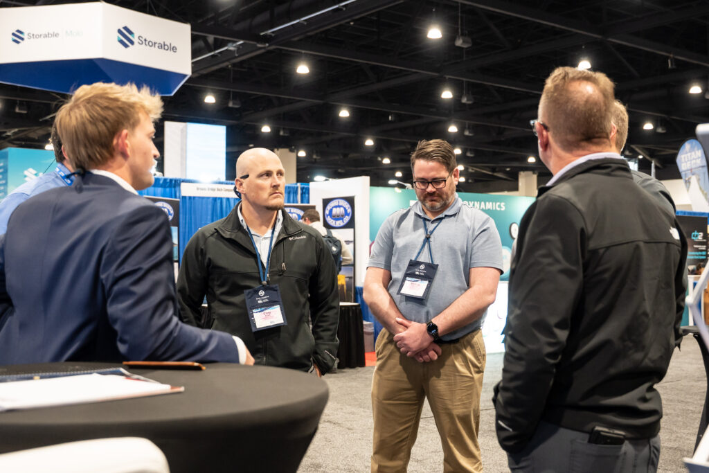 Three men engaged in conversation at a bustling trade show, surrounded by booths and attendees.