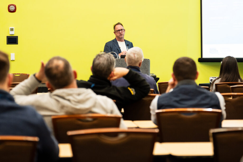 A bespectacled man presents at a conference, sharing insights with attendees while standing at a podium.