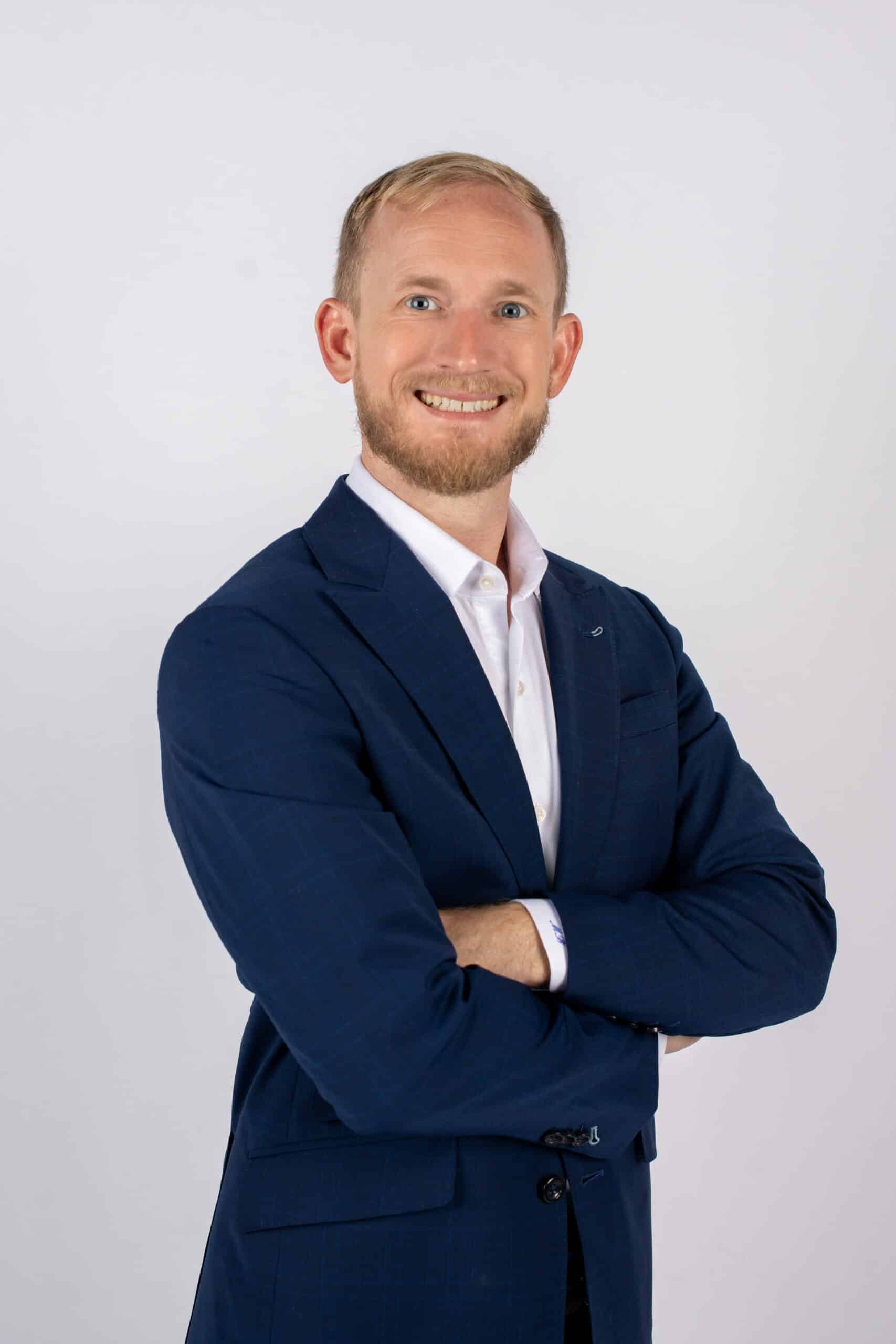 A man wearing a blue suit and a white shirt stands confidently, showcasing a professional appearance.