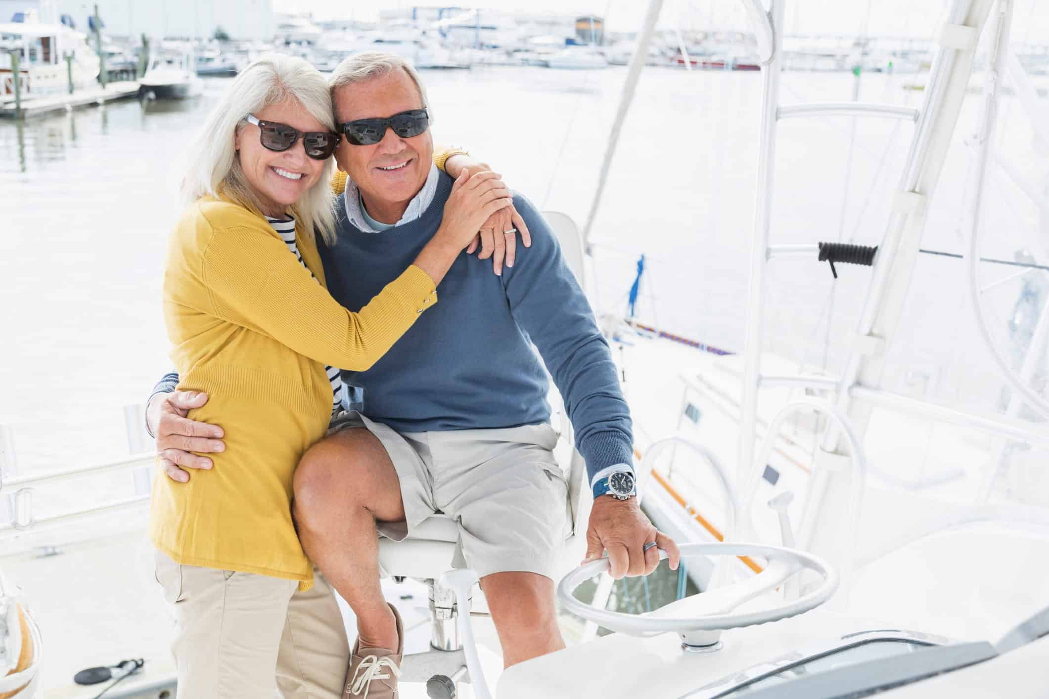 A happy, retired couple on their luxury boat, planning to spend the day on the water, sailing. The senior man is the captain, with one hand on the steering wheel and the other arm around his wife. She is hugging him and they are both smiling and looking forward.